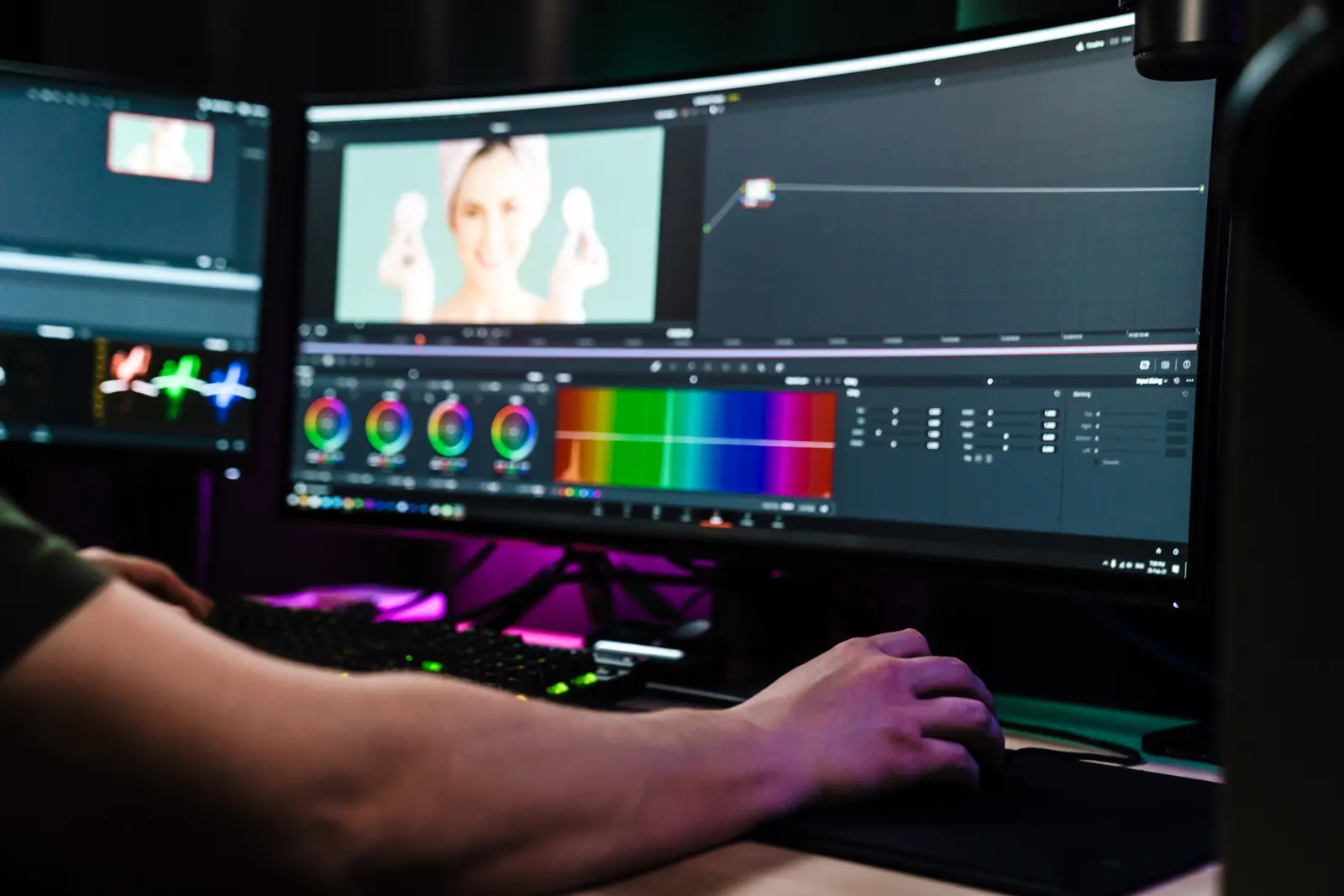 Video editor at a multi-monitor workstation with color-grading wheels visible on screen