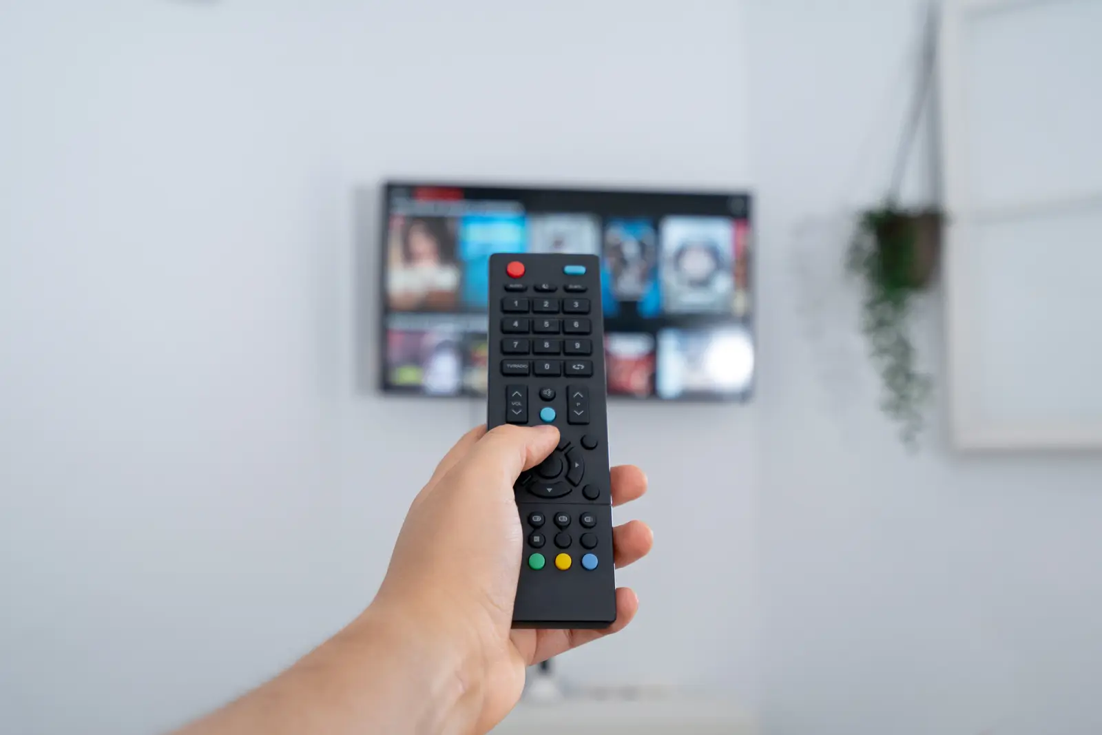 Hand holding TV remote pointing at a wall-mounted TV showing a streaming app menu